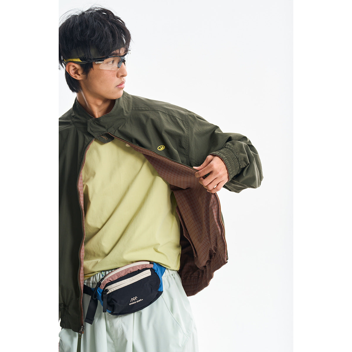 Model opening a dark olive jacket to show its brown plaid lining, over a pale yellow shirt.