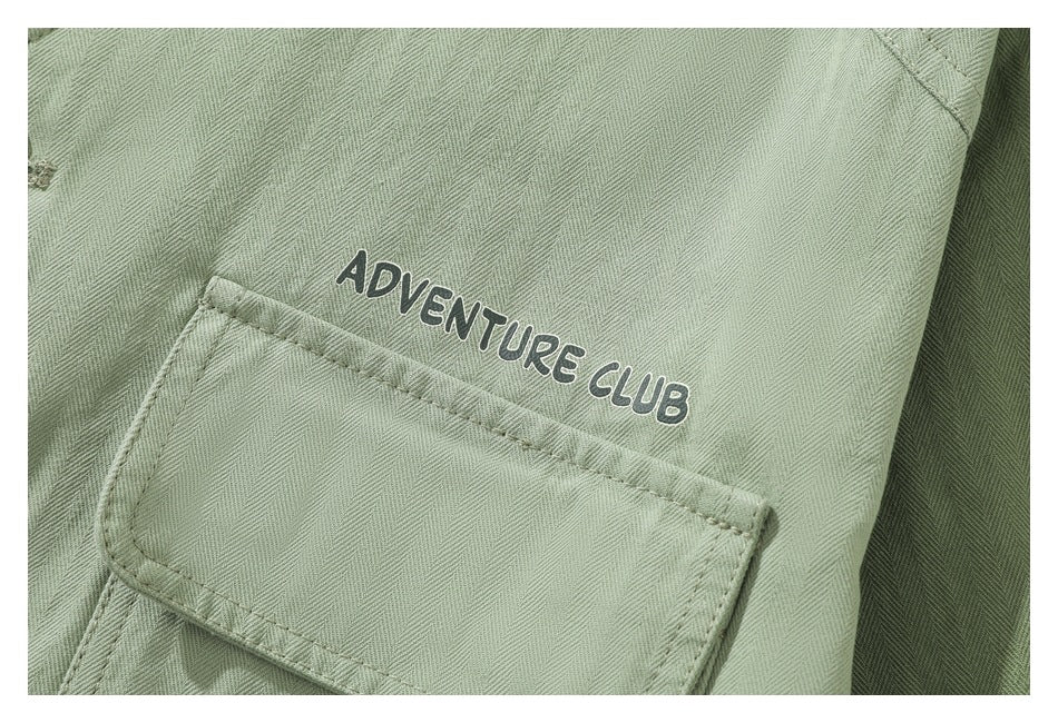 Close-up of a sage-green herringbone garment with a flap pocket and “Adventure Club” lettering.