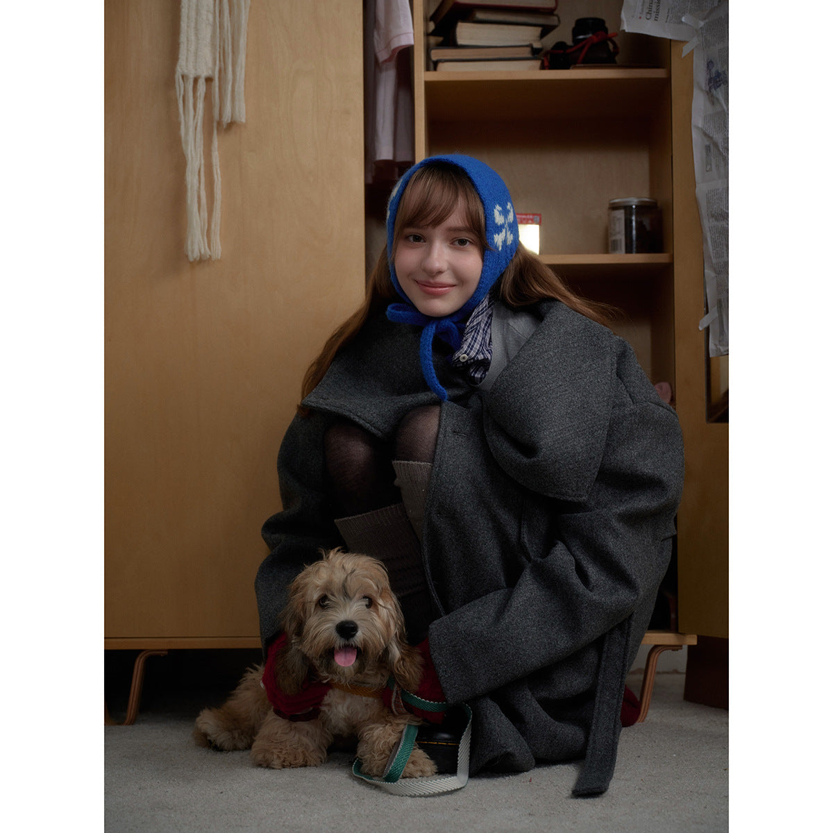 A woman crouches in an oversized charcoal-gray coat and blue patterned bonnet beside a small dog.