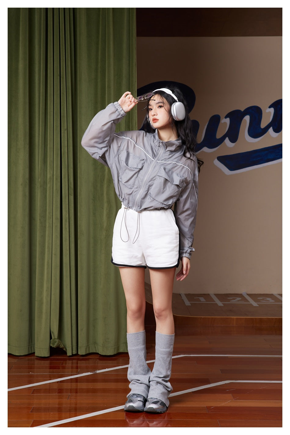 Model in cropped gray jacket with flap pockets and drawstring hem, raising one arm, with white shorts and gray leg warmers