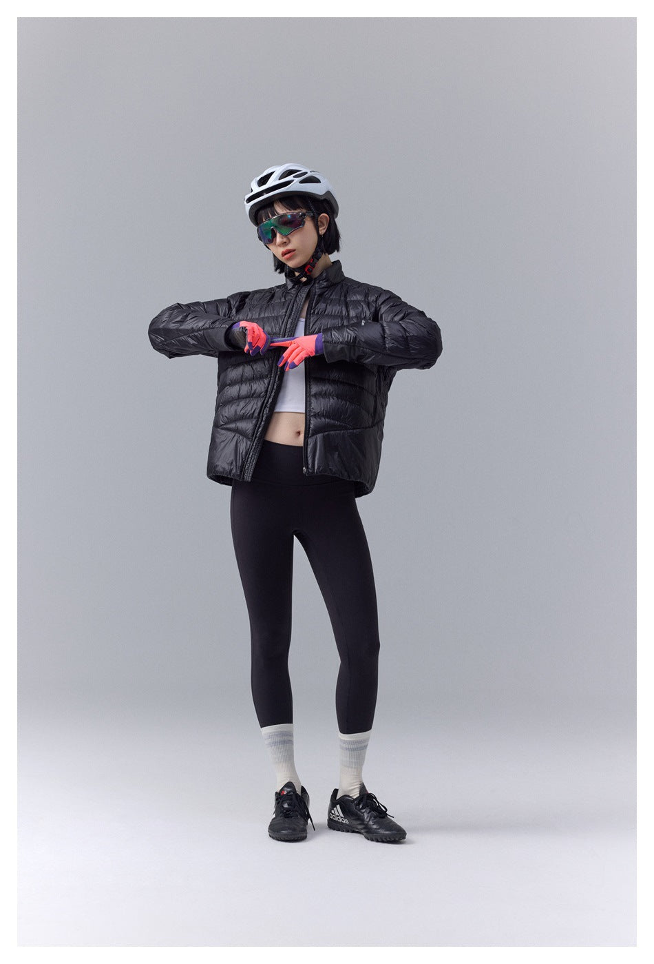 Full-length model wearing a black quilted jacket and cycling leggings, adjusting her gloves in a studio.