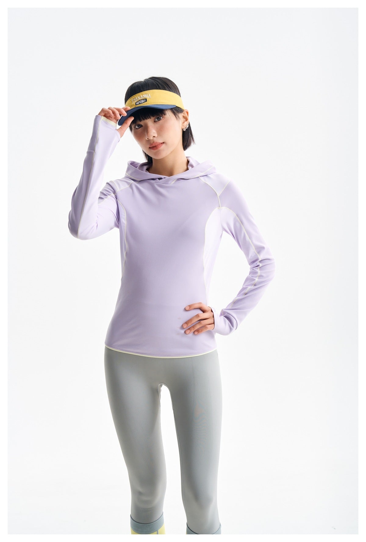 Model wearing a lavender hooded athletic top with pale contrast panels, gray leggings, and a yellow-and-navy visor.