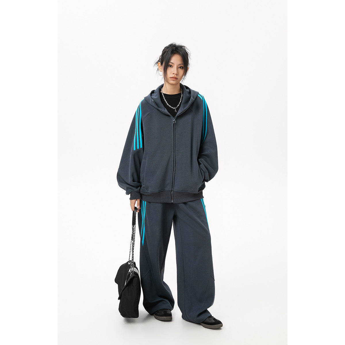 Model wearing a charcoal zip-up hoodie and loose pants with bright cyan stripes, carrying a black bag.