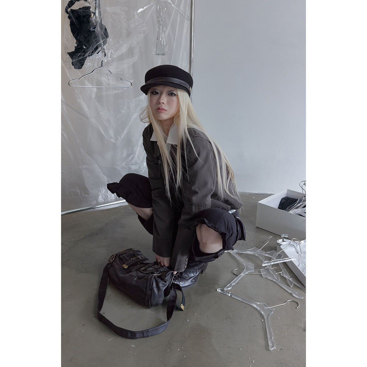 Crouching model wearing a charcoal jacket and black ruffled trousers with a black cap and boots.