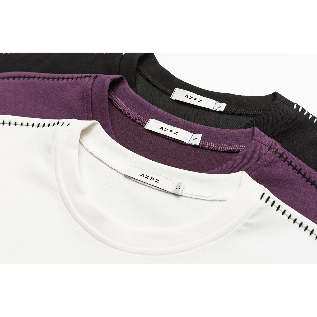 Close-up of white, plum, and black crewneck T-shirts with visible neck labels and black decorative shoulder stitching.