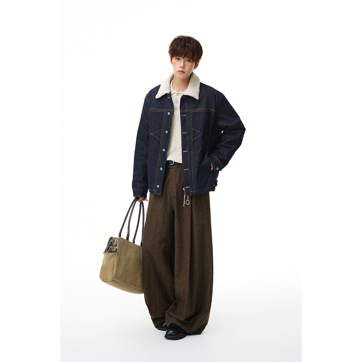 Full-length model wearing a dark indigo denim jacket with a cream sherpa collar, brown wide-leg trousers, and carrying a tan