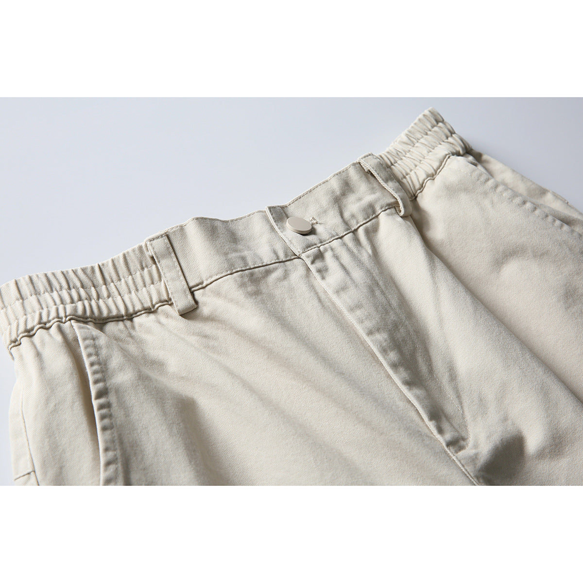 Close-up of light beige pants showing the elastic waistband, belt loop, button, and front closure.