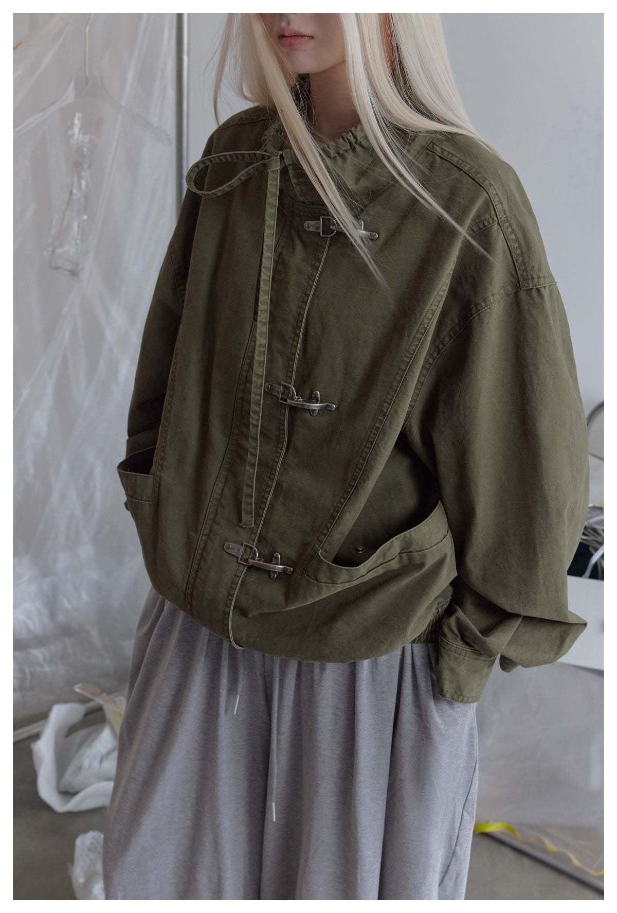 Close view of a model wearing an olive jacket with metal hook closures, roomy sleeves and large front pockets.