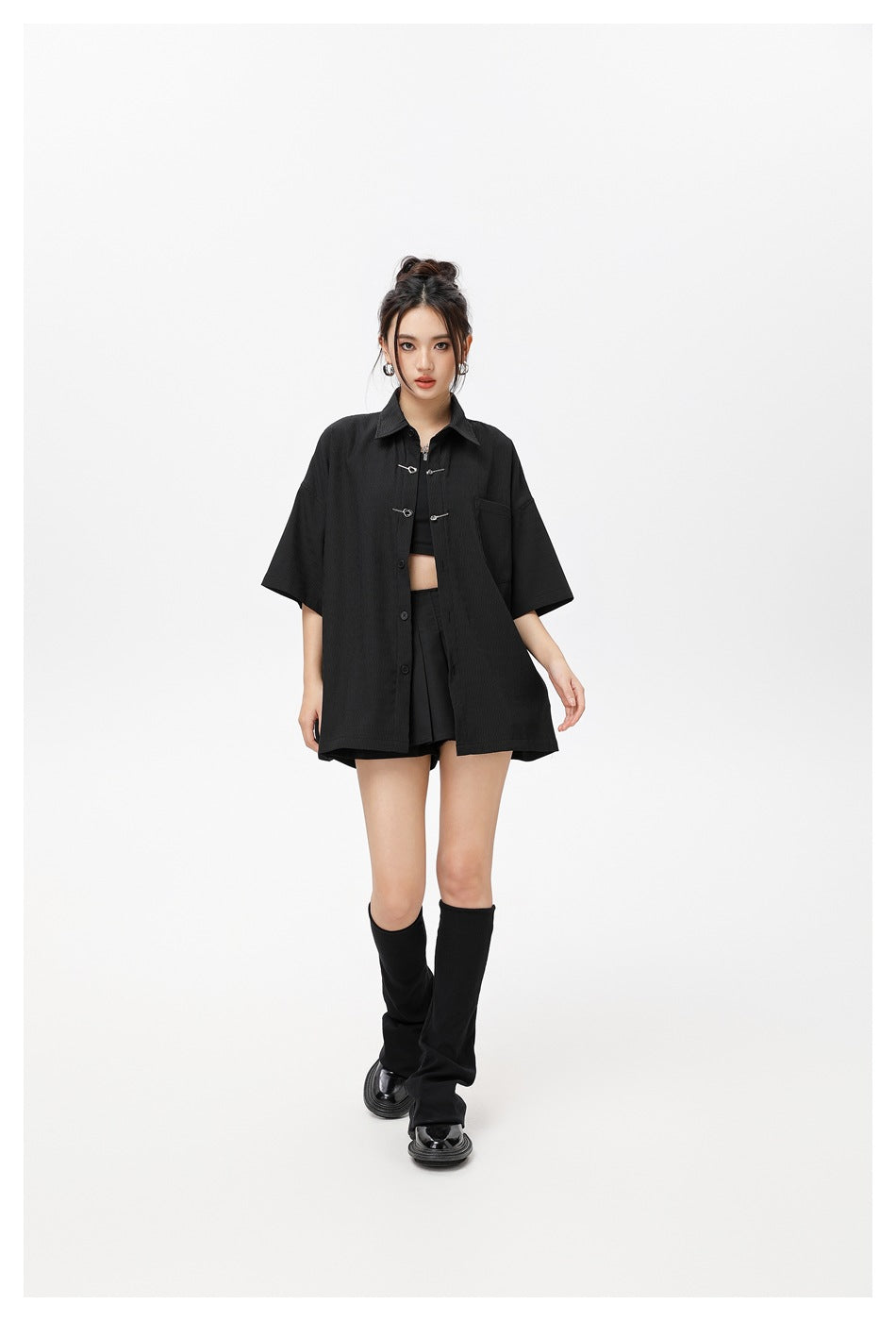 Model wearing the black oversized short-sleeve shirt open over a black crop top, pleated skirt and leg warmers