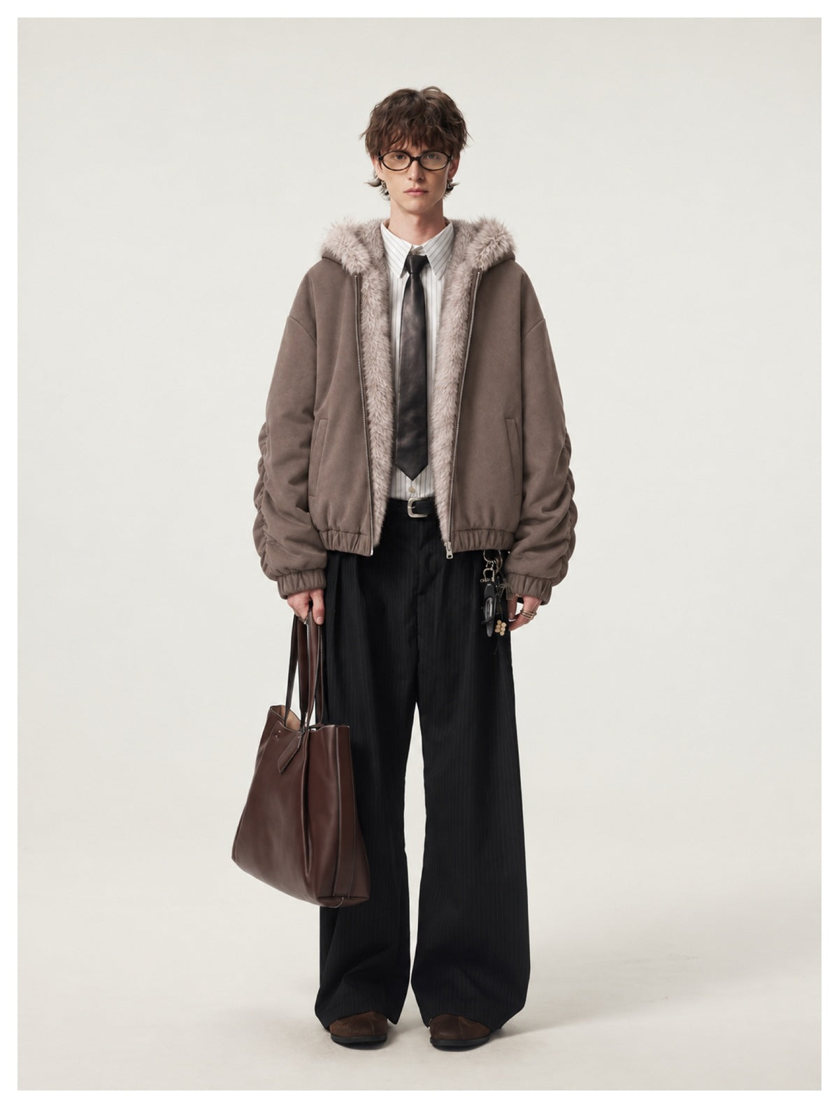 Full-length view of a man wearing a taupe faux-fur-trimmed hooded jacket with a shirt, tie, and wide black trousers.