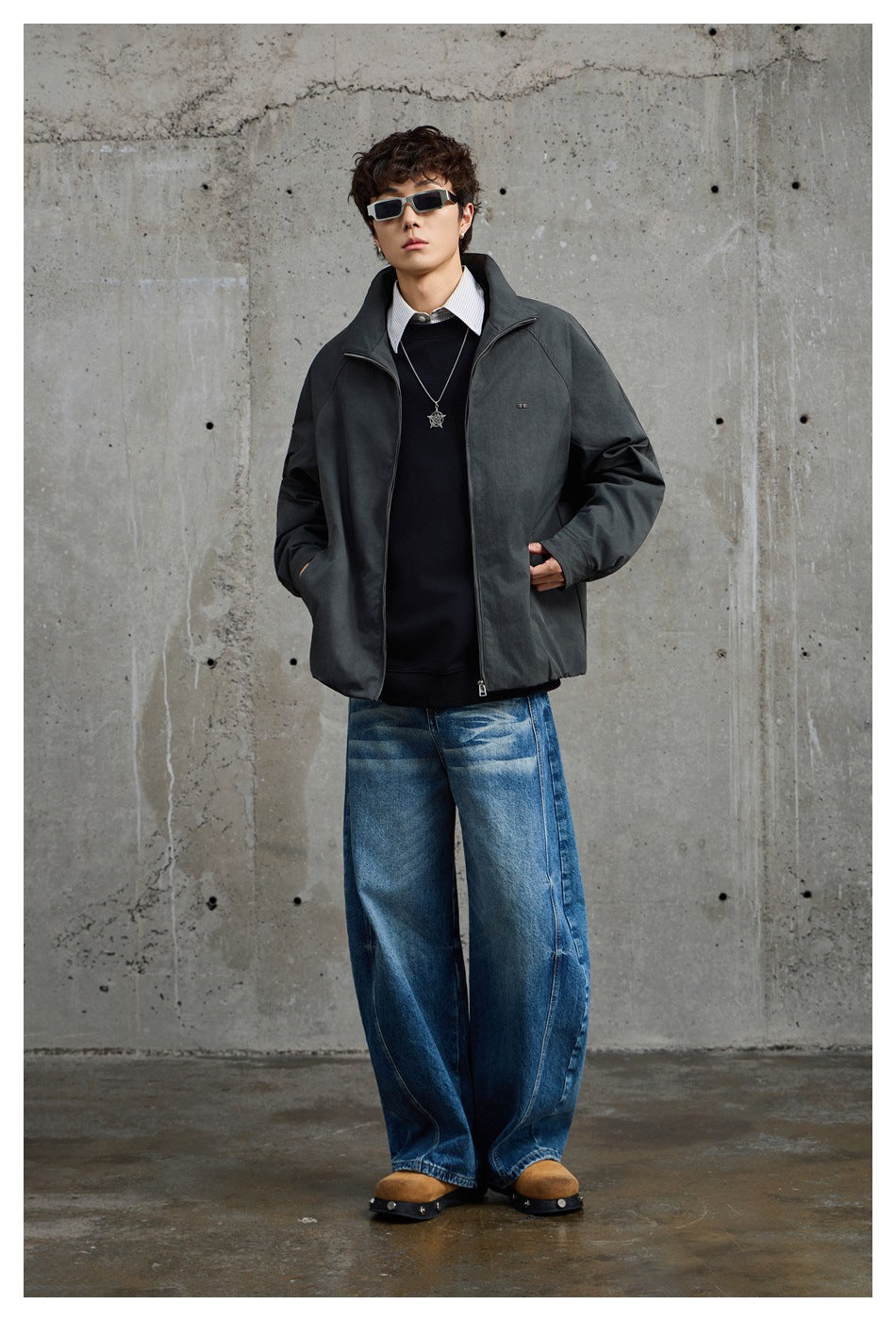Model wearing a charcoal zip-front jacket over a black sweater and white shirt, with loose blue jeans.