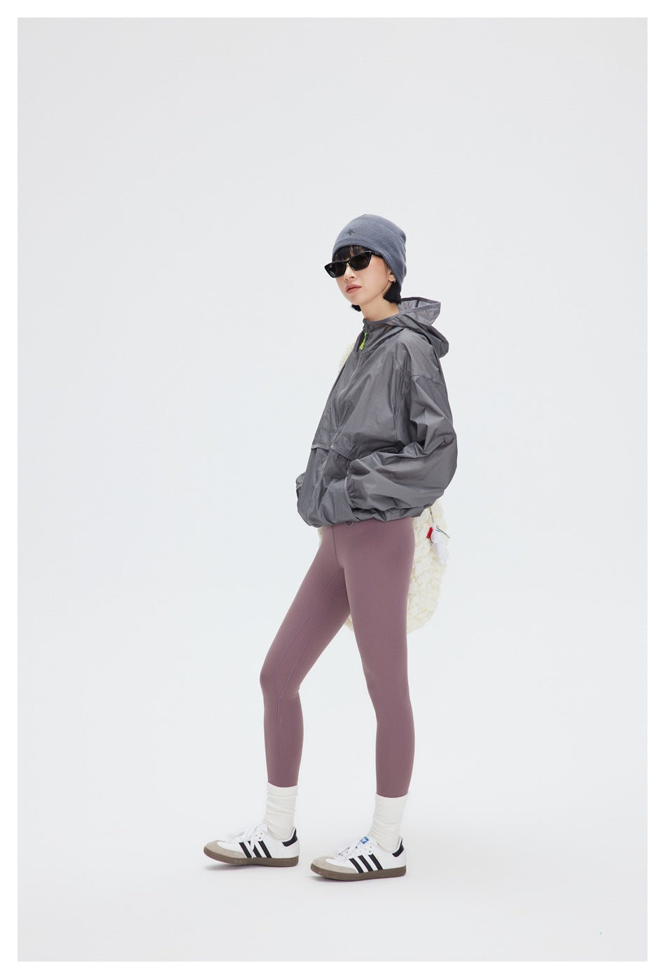 Model in a gray hooded windbreaker and mauve leggings, shown in a three-quarter pose with a textured white shoulder bag.