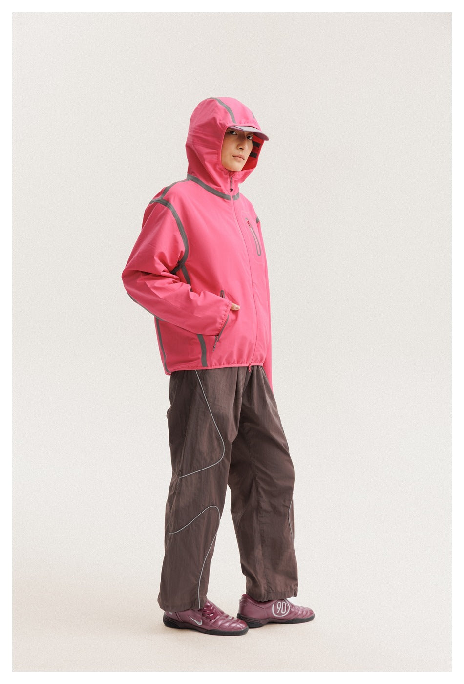 Model wearing a bright pink hooded jacket with gray trim and dark brown trousers, hood up, in a three-quarter view.