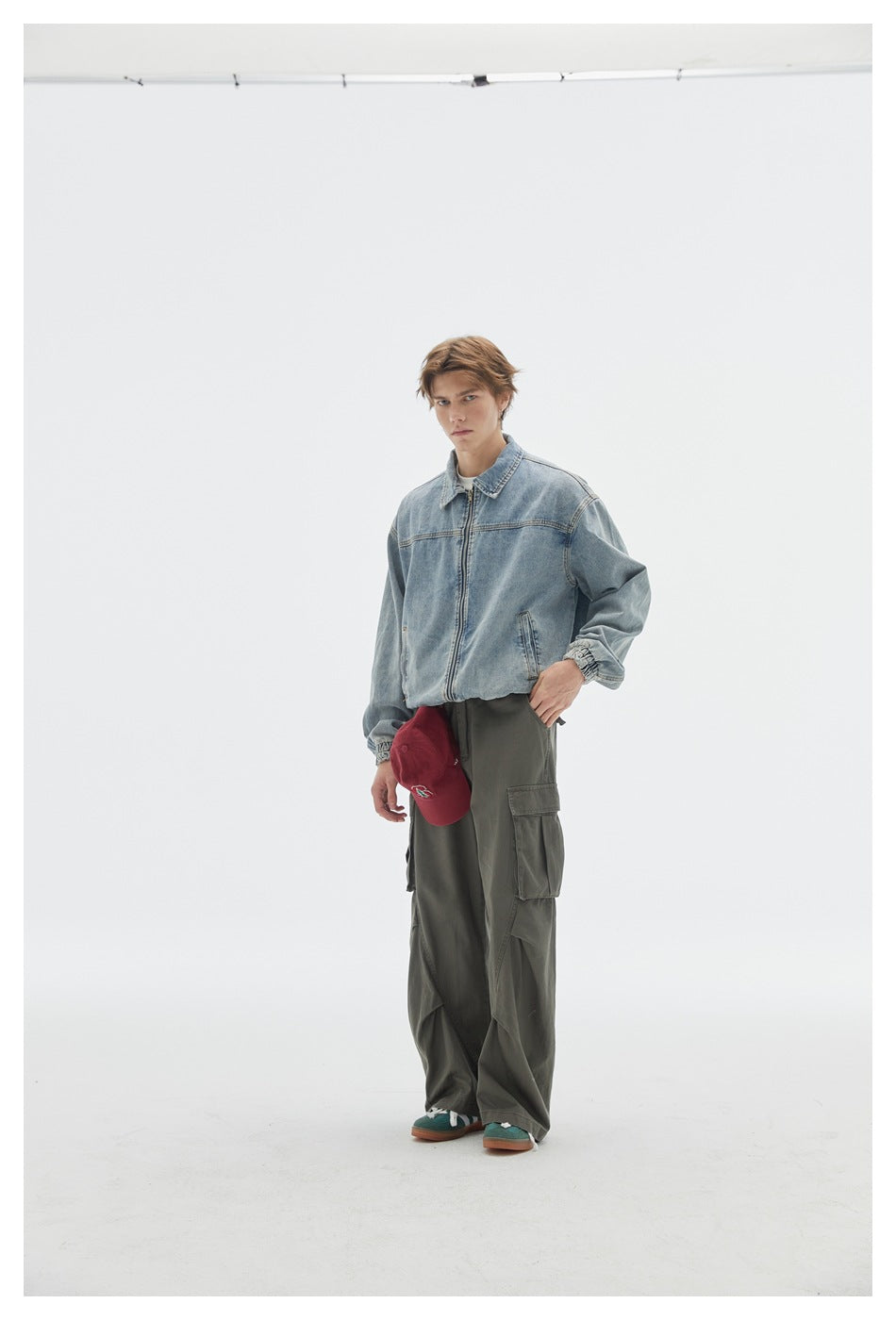 Model wearing a light blue zip-front denim jacket and loose olive cargo pants, holding a red cap.