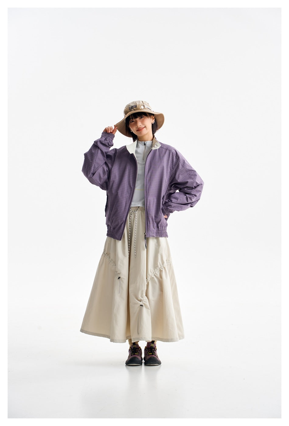 Full-length front view of a model wearing a purple plaid jacket with a cream skirt and hiking shoes.