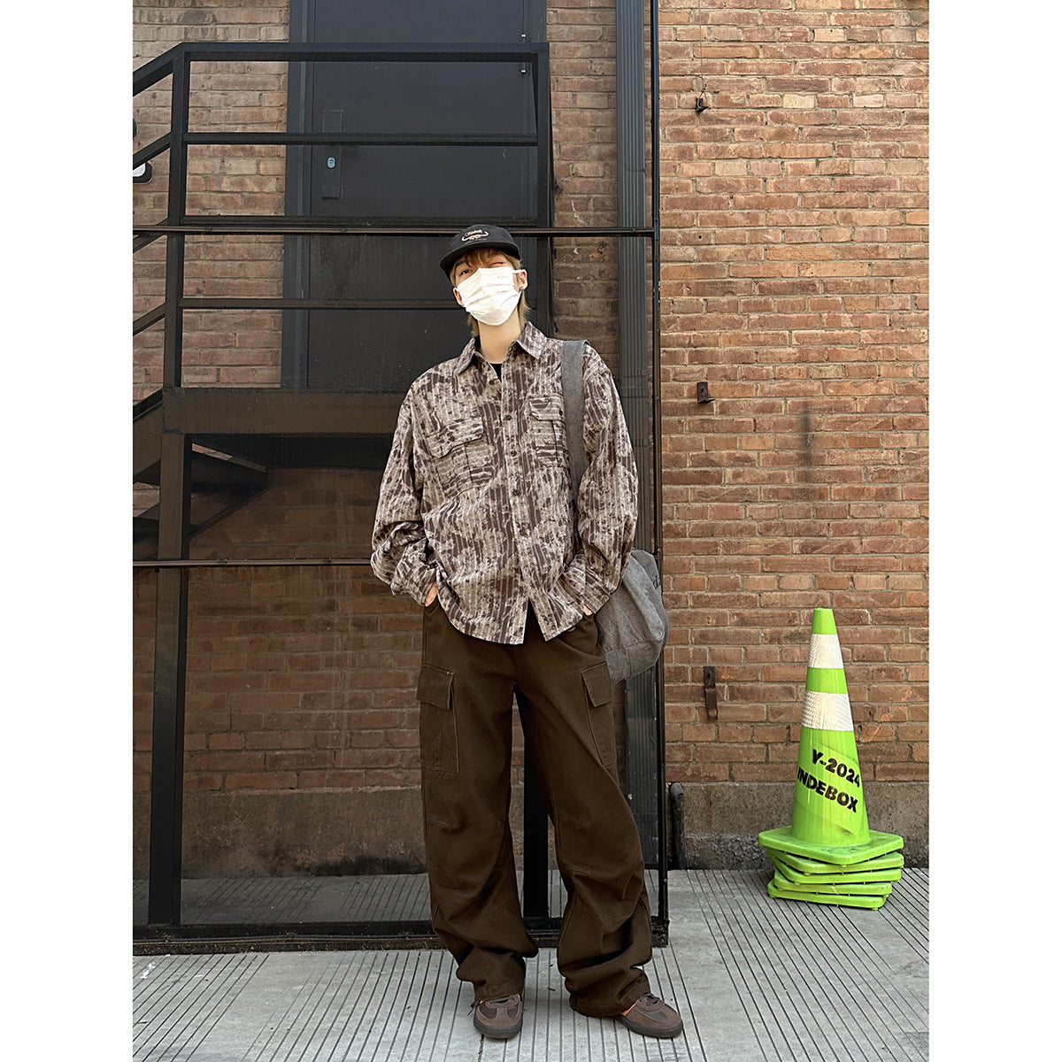 Model wearing a brown and beige patterned button-up shirt with dark brown cargo pants and a gray shoulder bag.