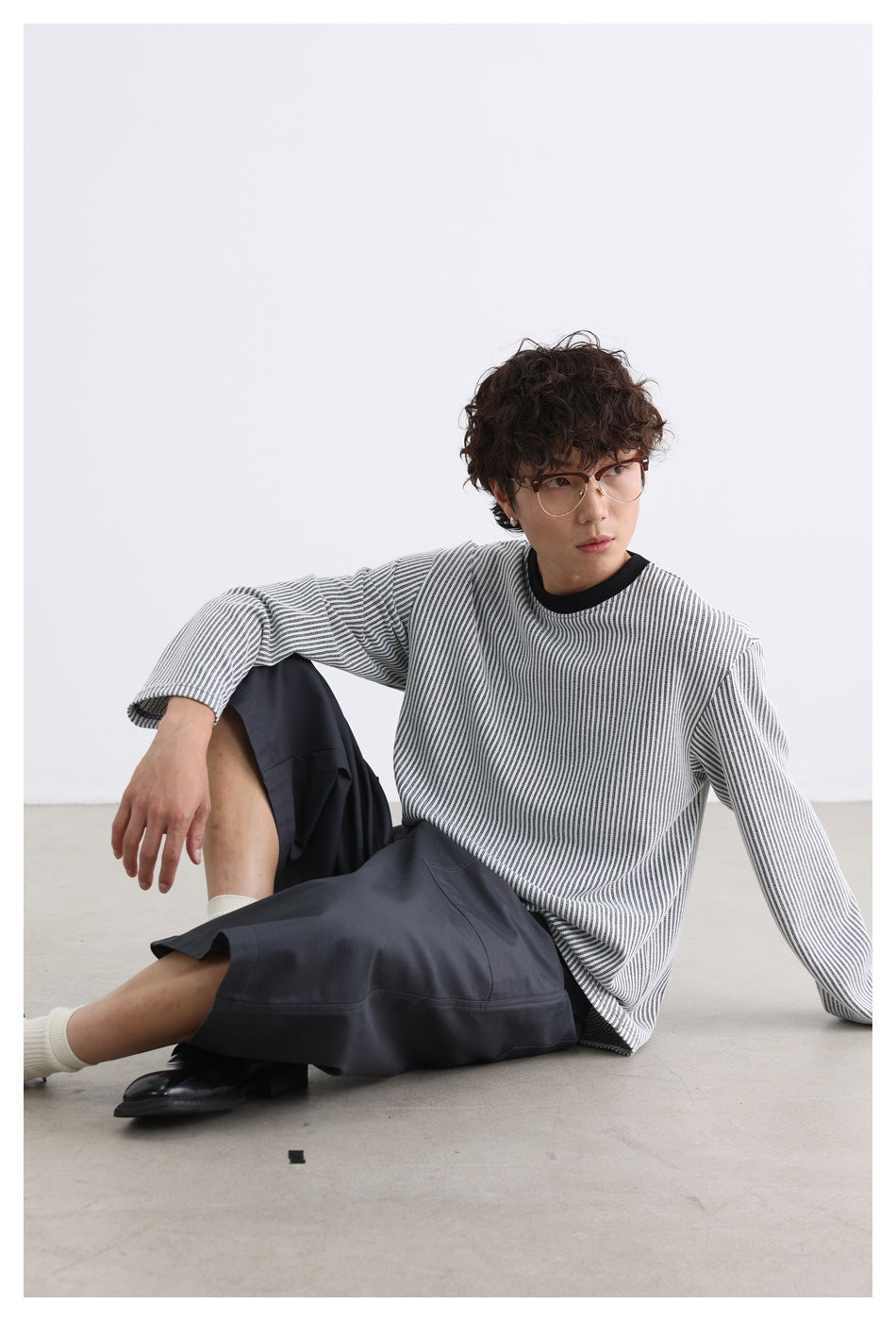 Model seated on the floor wearing a gray-white striped long-sleeve crew-neck top with gray pants and black shoes