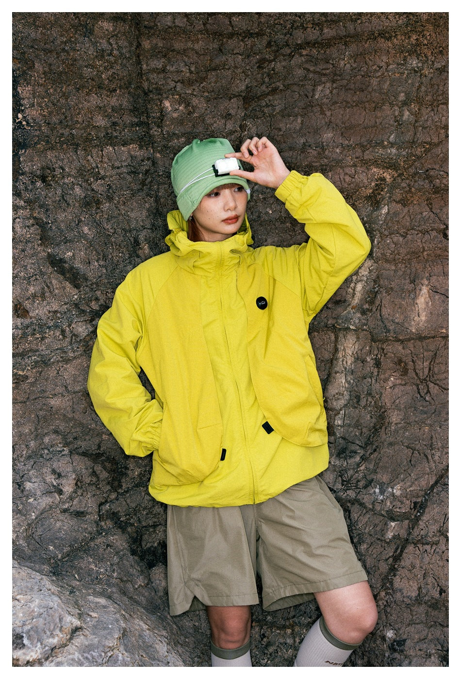 Close view of a model wearing a bright yellow hooded jacket with large front pockets and taupe shorts.