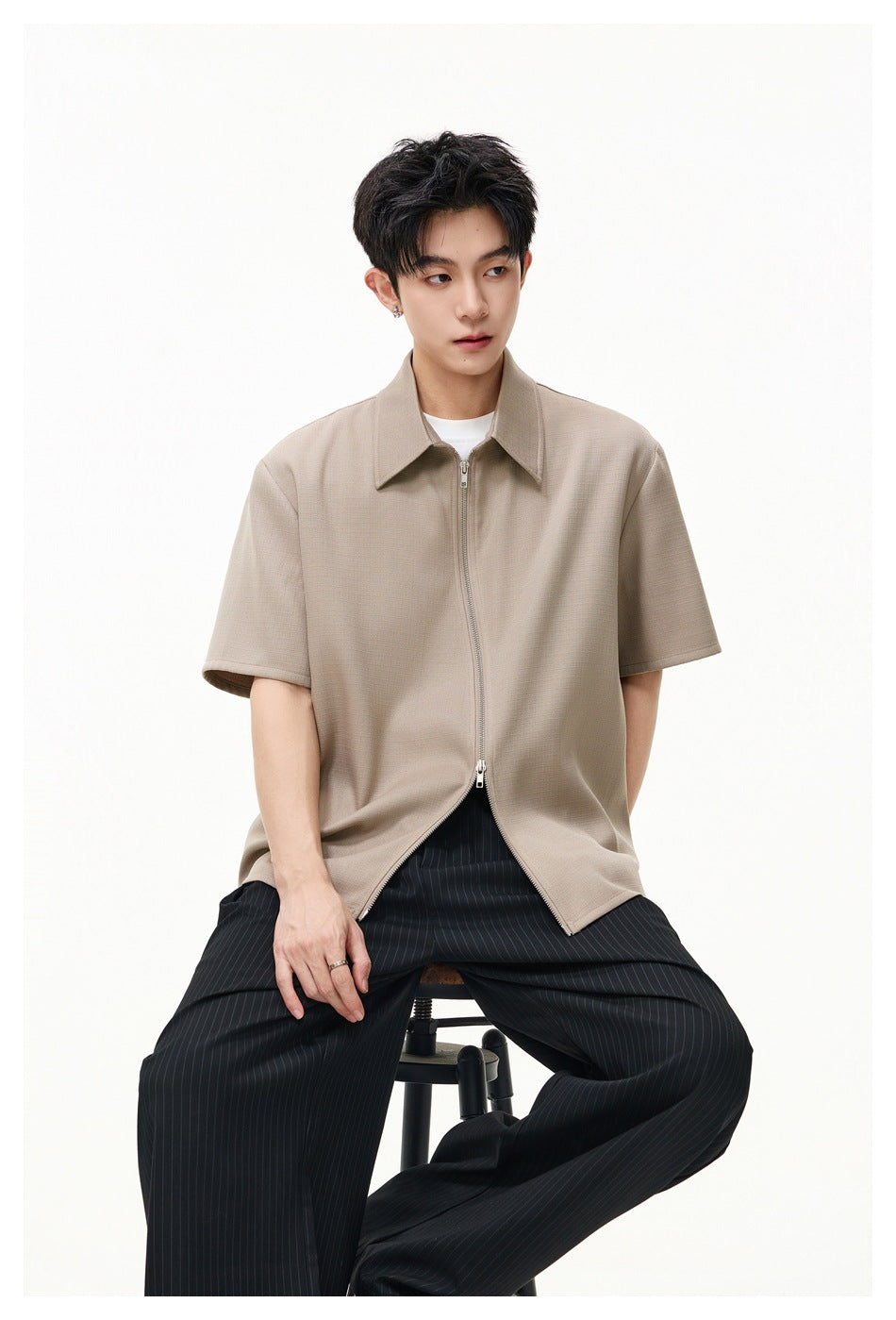 Model seated on a stool wearing a tan zip-front short-sleeve shirt with white tee and pinstripe trousers