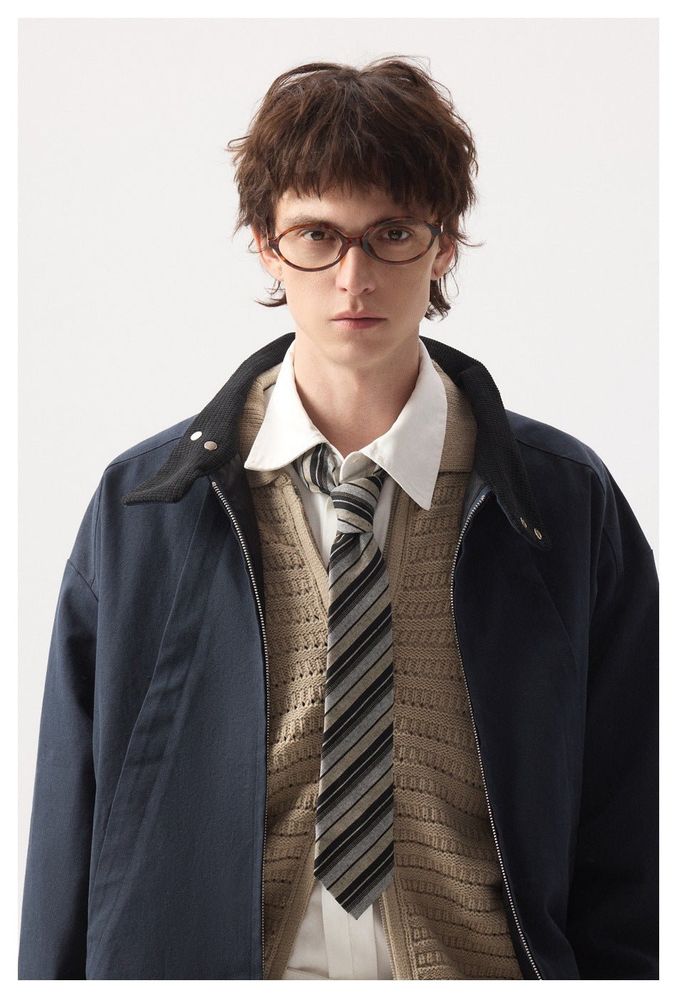 Close view of a model wearing an open navy jacket over a knit vest, white shirt and striped tie.