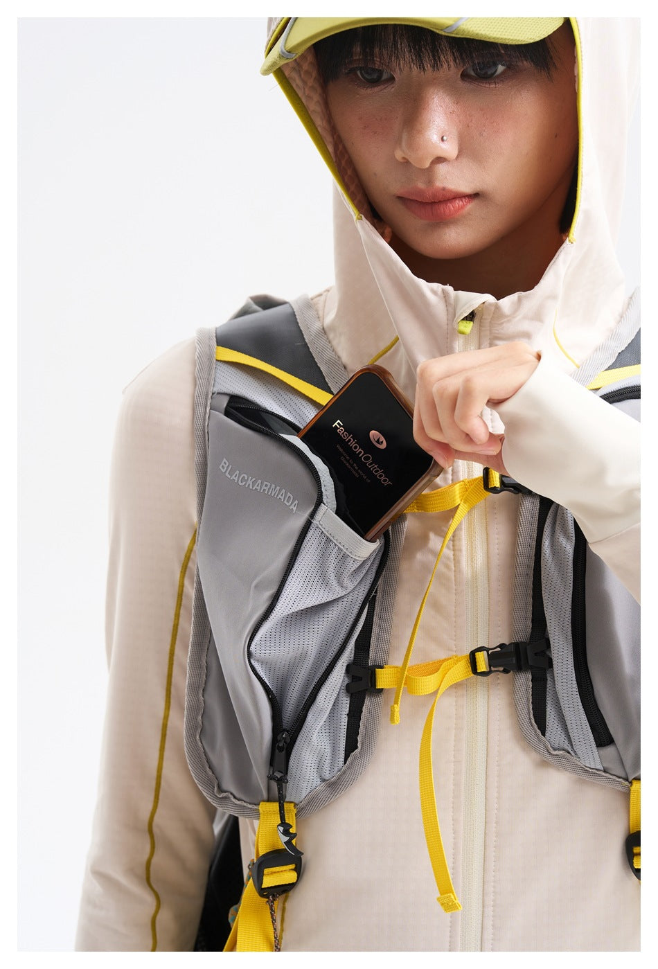 Close-up of a model wearing a cream hooded top and gray chest pack with yellow straps, placing a phone into a zip pocket.