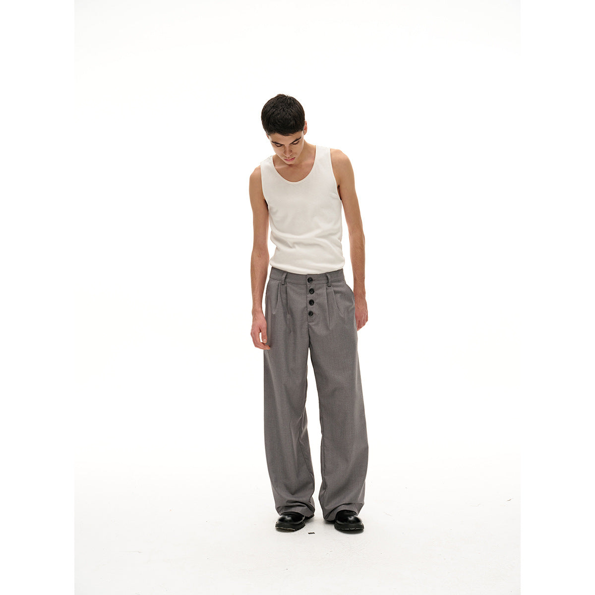 Model looking down in a white tank top and loose gray pleated trousers with a four-button front closure.