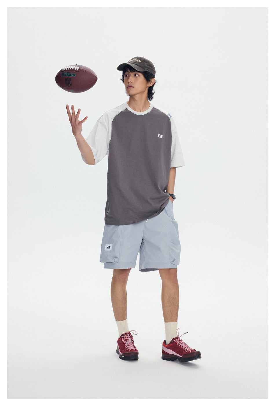 Full-length model wearing a charcoal-and-light-gray raglan T-shirt and pale blue shorts while tossing a football.