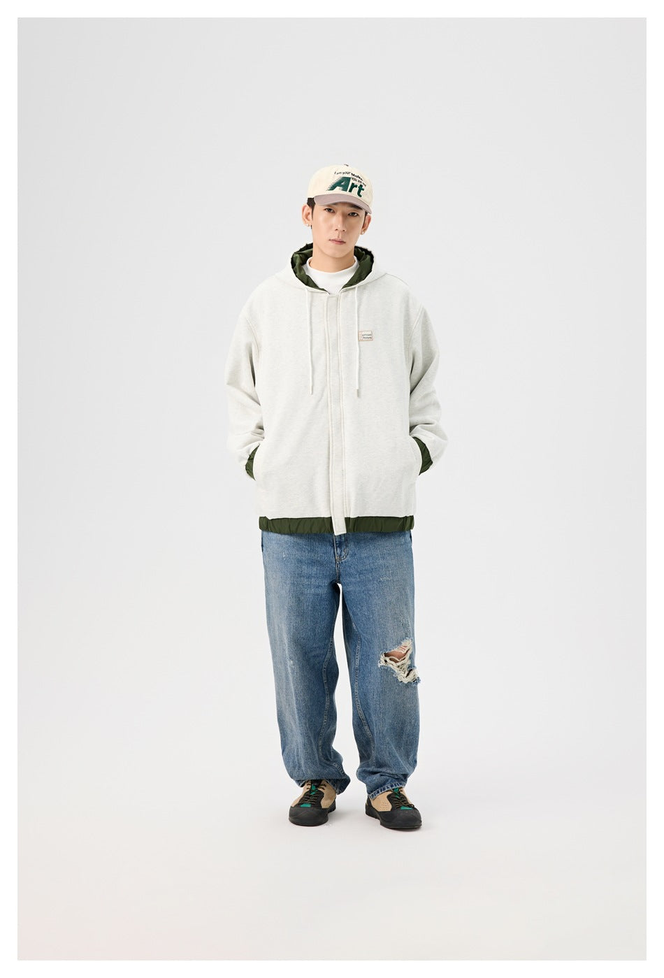 Man in gray hooded zip jacket with green trim at hem, hands in pockets, with cap, ripped jeans and sneakers