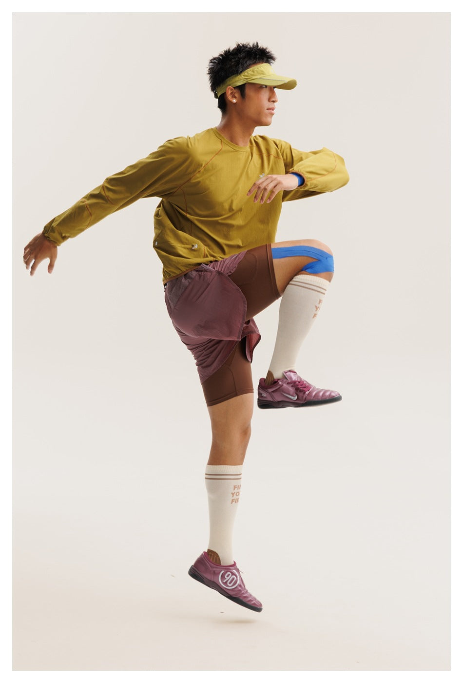 Model in a mustard-yellow long-sleeve top and mauve shorts, lifting one knee in an athletic pose.
