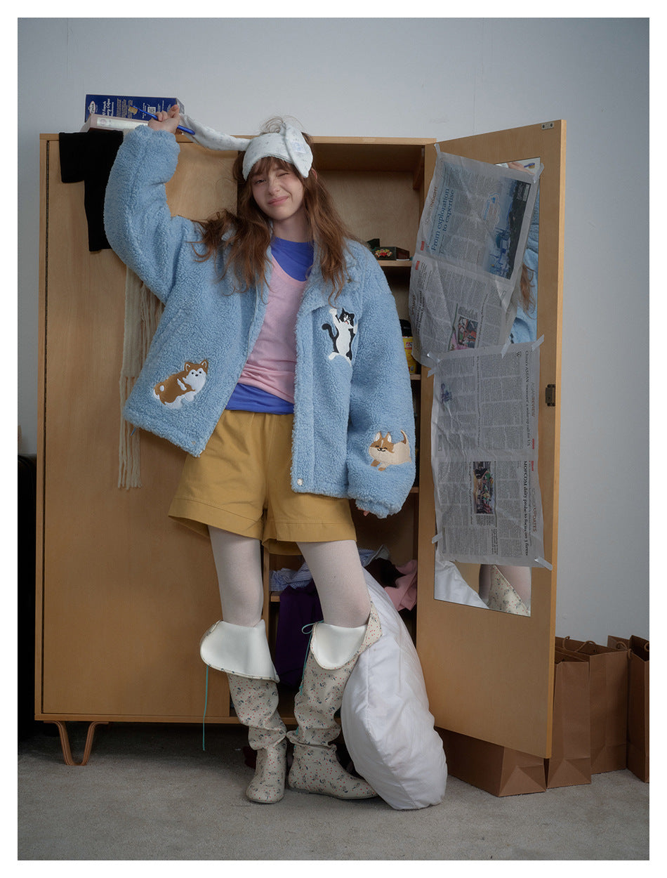 A model wearing a light blue plush jacket with colorful animal appliqués, shown with mustard shorts.