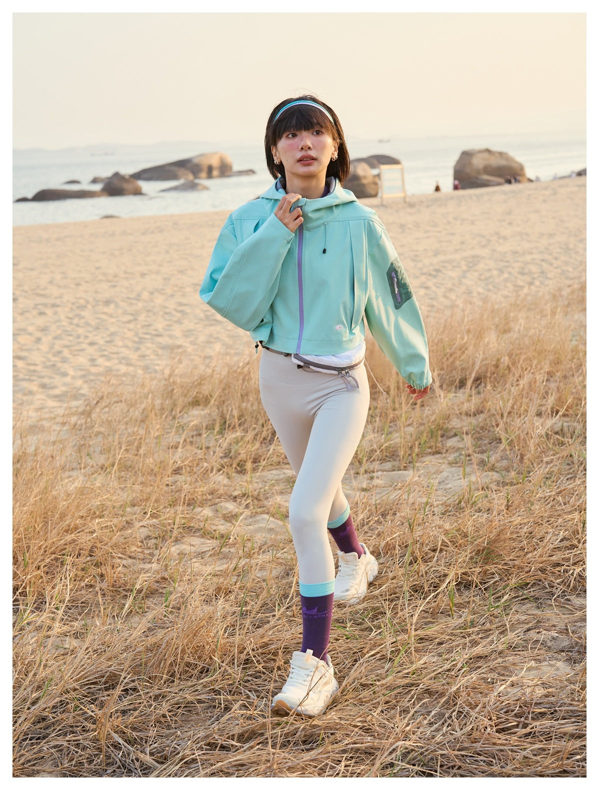 Model wearing a mint cropped hooded jacket with a purple zipper, light leggings and purple socks on a beach.