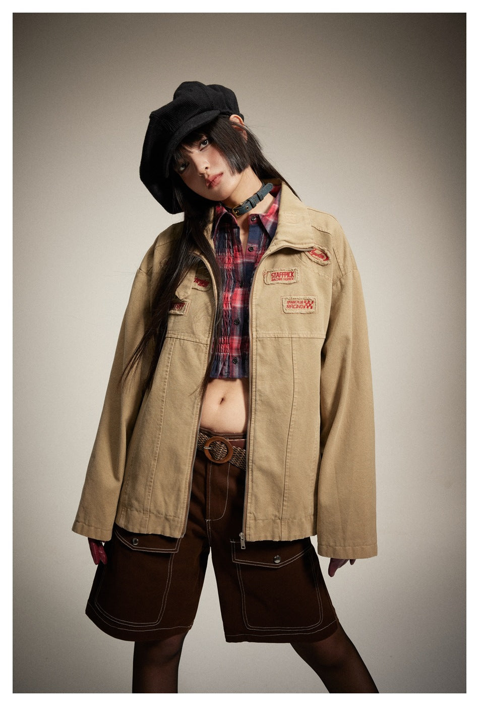 Model wearing a tan embroidered zip jacket over a red plaid crop top and brown shorts.