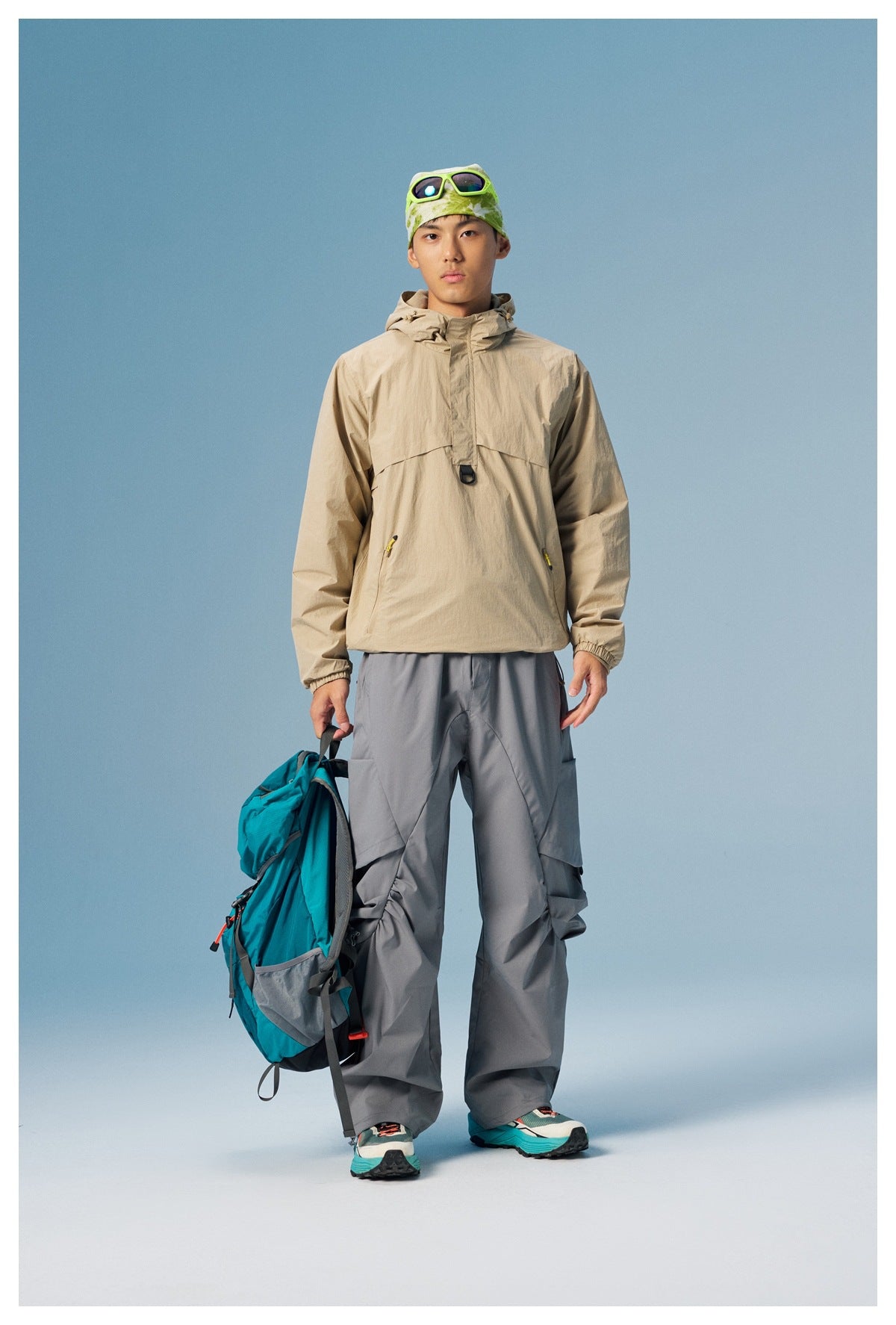 Model wearing a beige hooded pullover and gray cargo pants, holding a teal backpack.