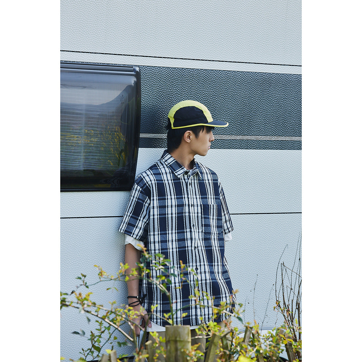 Side view of a model wearing an oversized navy and white plaid short-sleeve shirt and neon yellow cap.