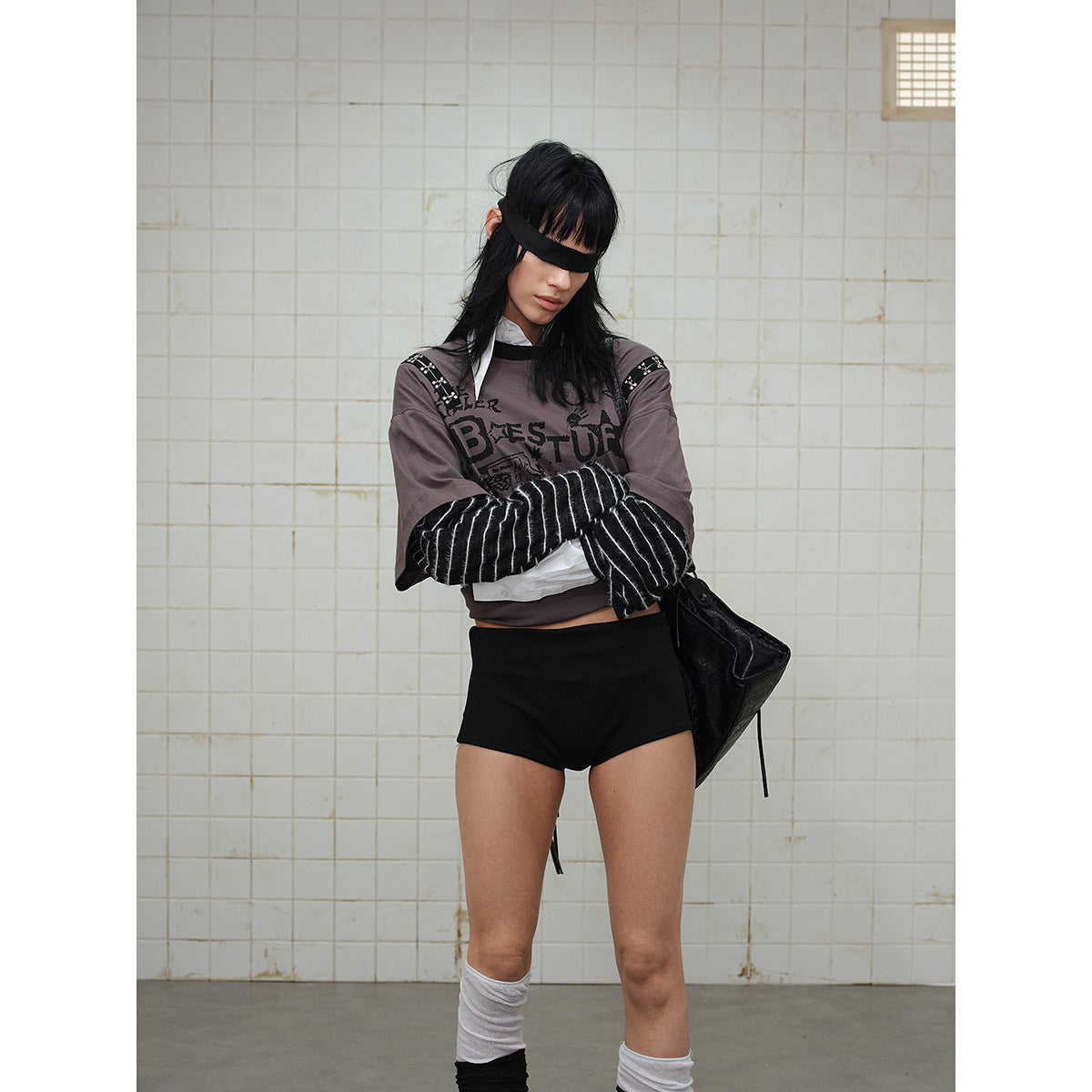 Model wearing a gray graphic top with striped sleeves and black shorts, posed with arms crossed.