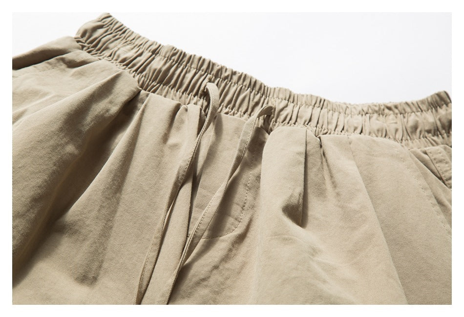 Close-up of tan shorts showing the gathered elastic waistband, drawstring, front pleats and pocket opening.