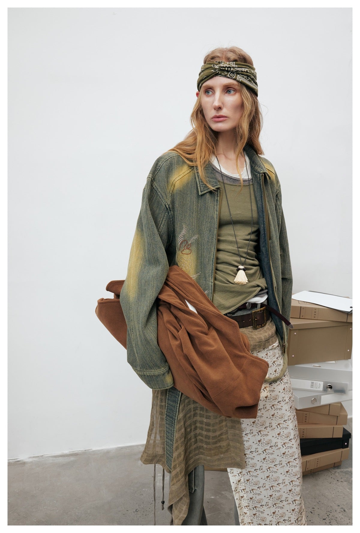 Model wearing an olive distressed embroidered jacket over layered tops, with a patterned skirt and brown bag.