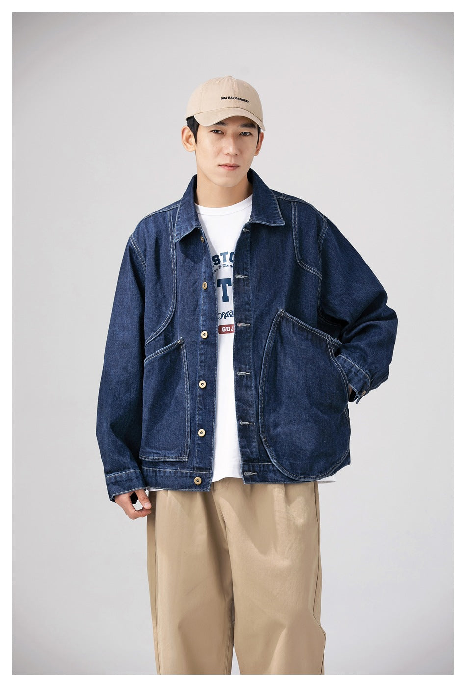 Model wearing a dark indigo denim work jacket with oversized patch pockets and tan buttons, paired with beige trousers and a