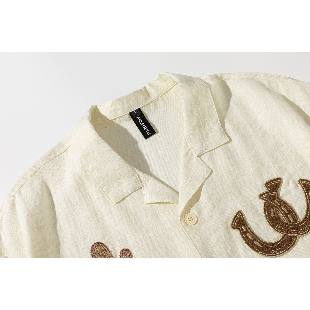 Close-up of an ivory collared shirt with a black neck label and brown embroidered horseshoe motifs.