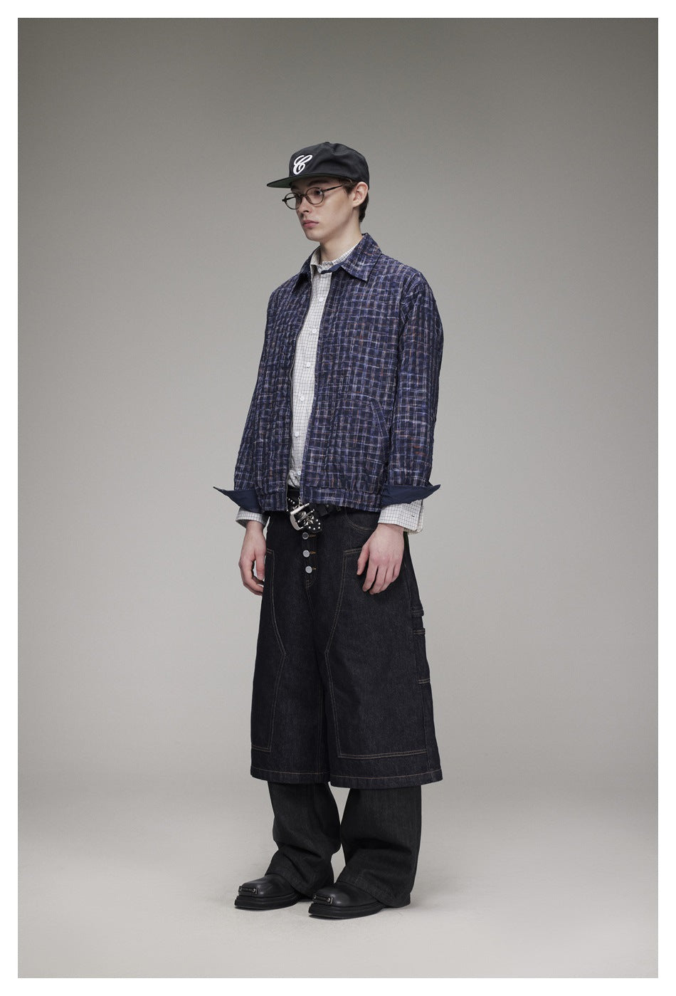 Model wearing a dark navy plaid jacket over a light checked shirt, with black wide shorts and a black cap; front view.