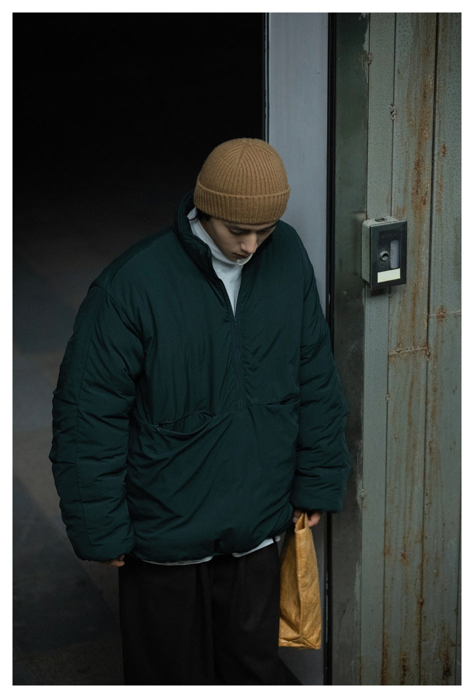Man in a dark green half-zip padded jacket and tan beanie looking down, standing by a rusty metal door