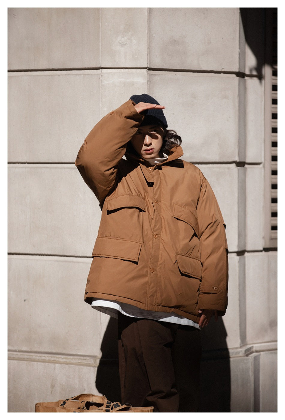 Model in an oversized khaki padded jacket with a navy beanie and brown trousers, hand raised to shade eyes