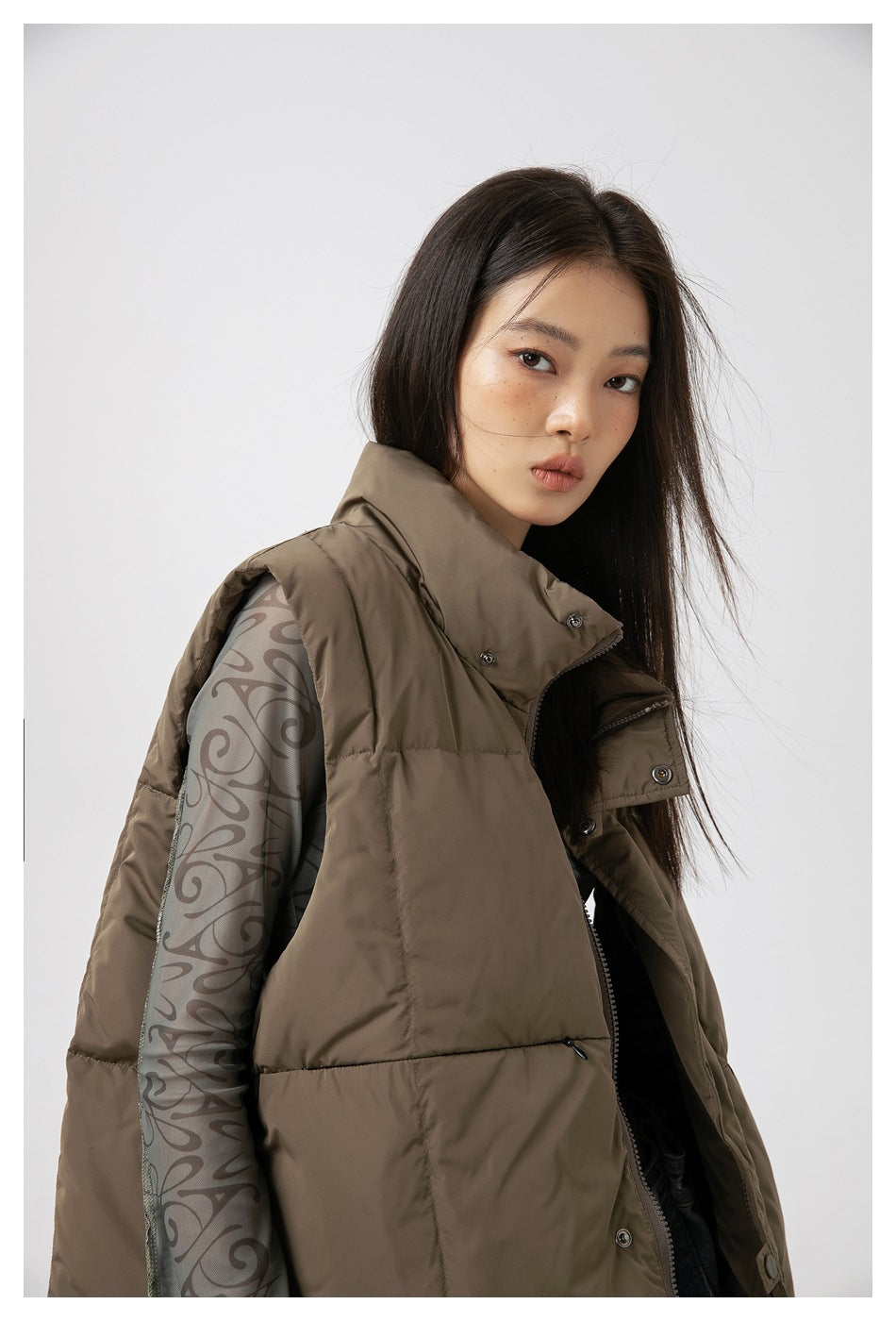 Close view of a model in a taupe quilted vest with a high collar and snap front, over a patterned gray top.