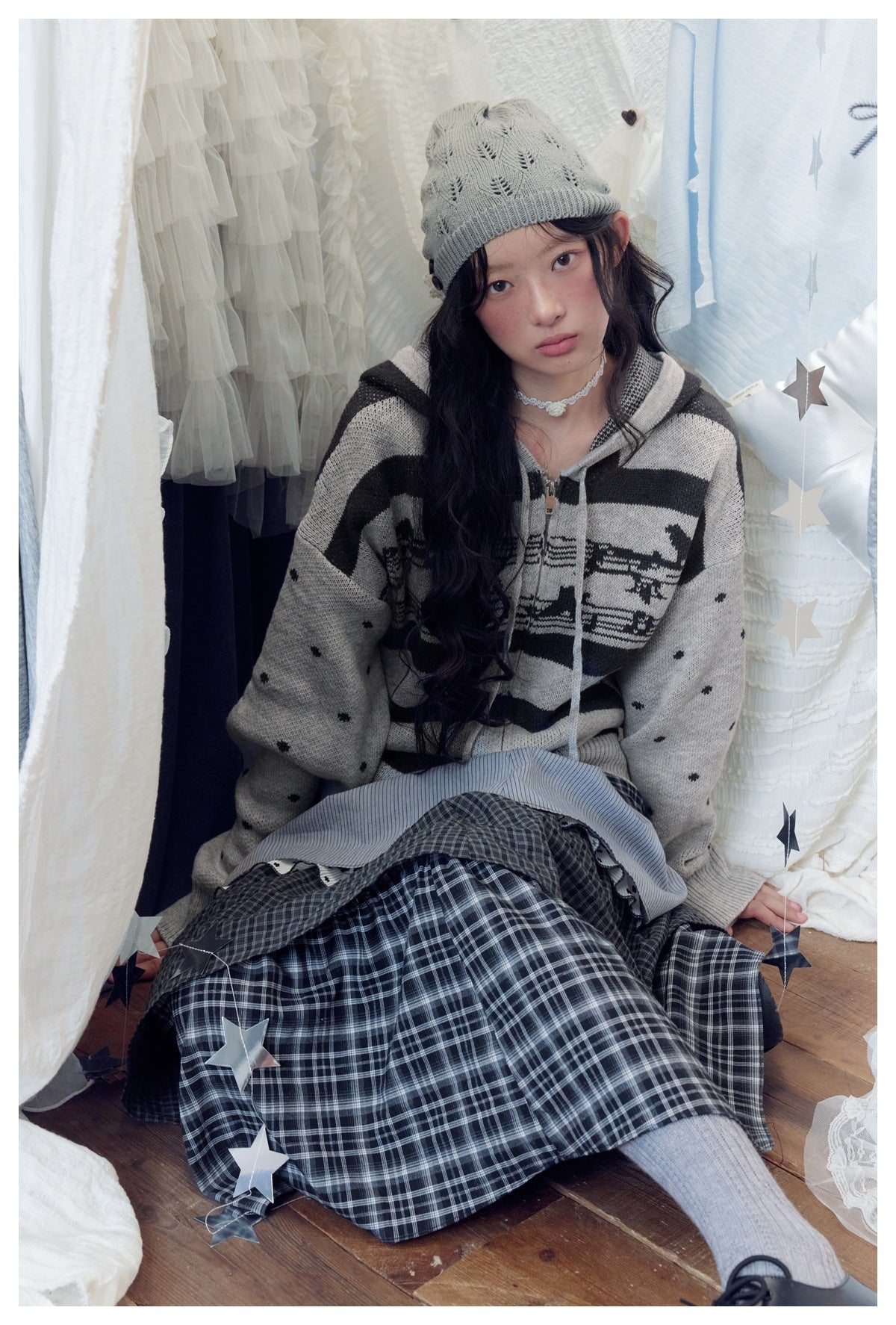 Seated model wearing a gray and black animal-pattern hooded sweater and a dark plaid skirt.