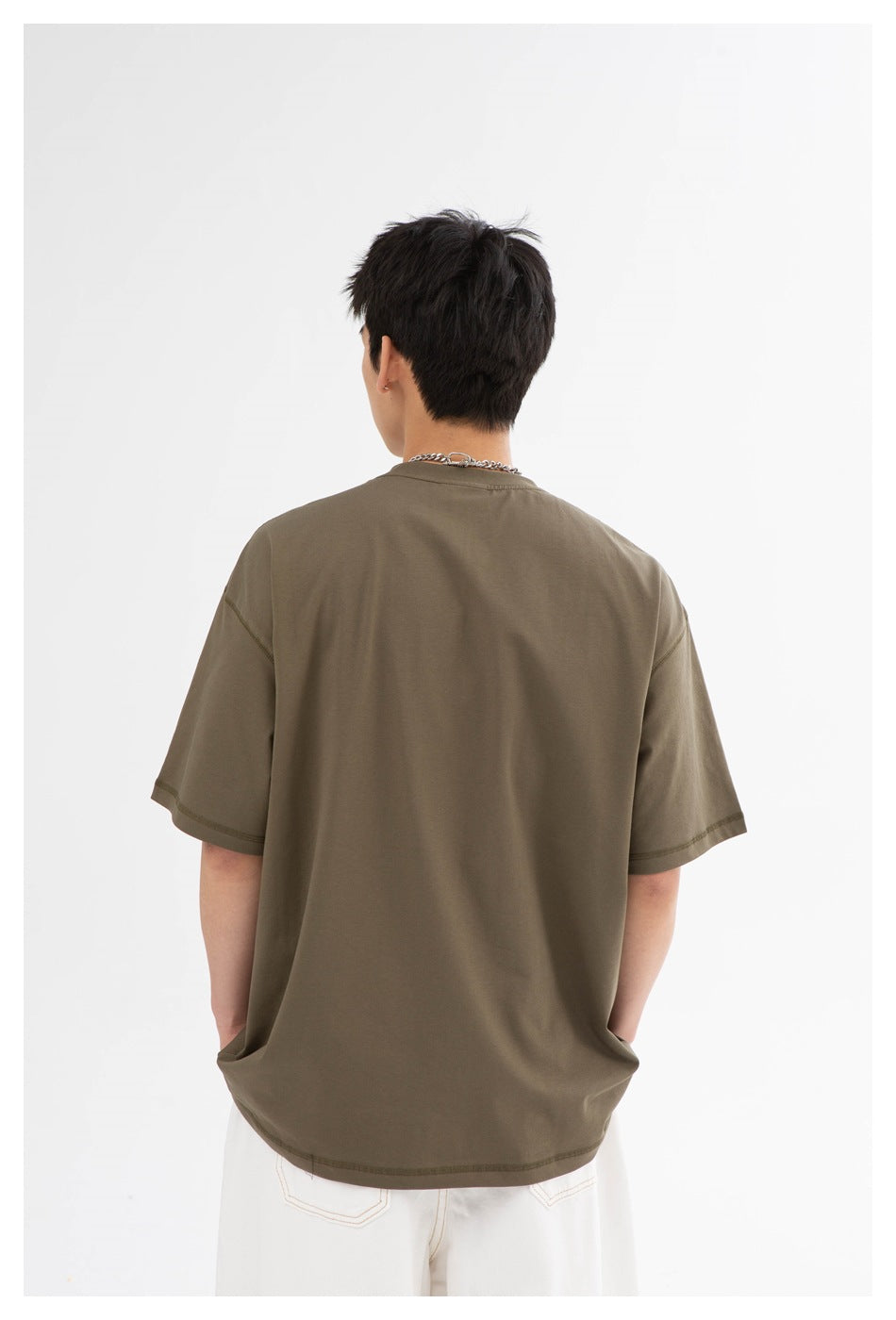 Back view of a model wearing a loose olive-brown short-sleeve T-shirt with a crew neck and white pants.