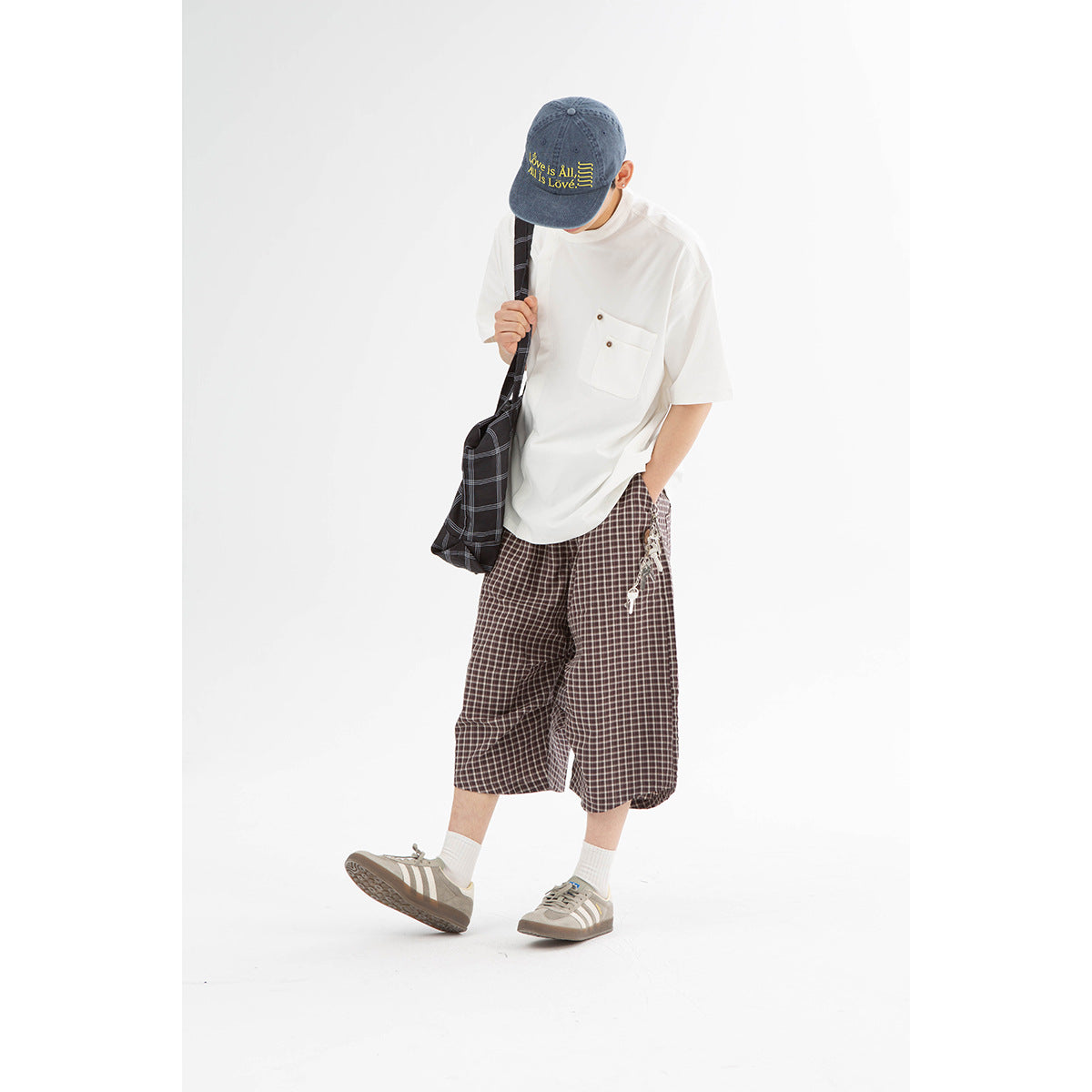 Model wearing a white short-sleeve shirt with a buttoned chest pocket and brown plaid shorts, carrying a dark plaid bag.