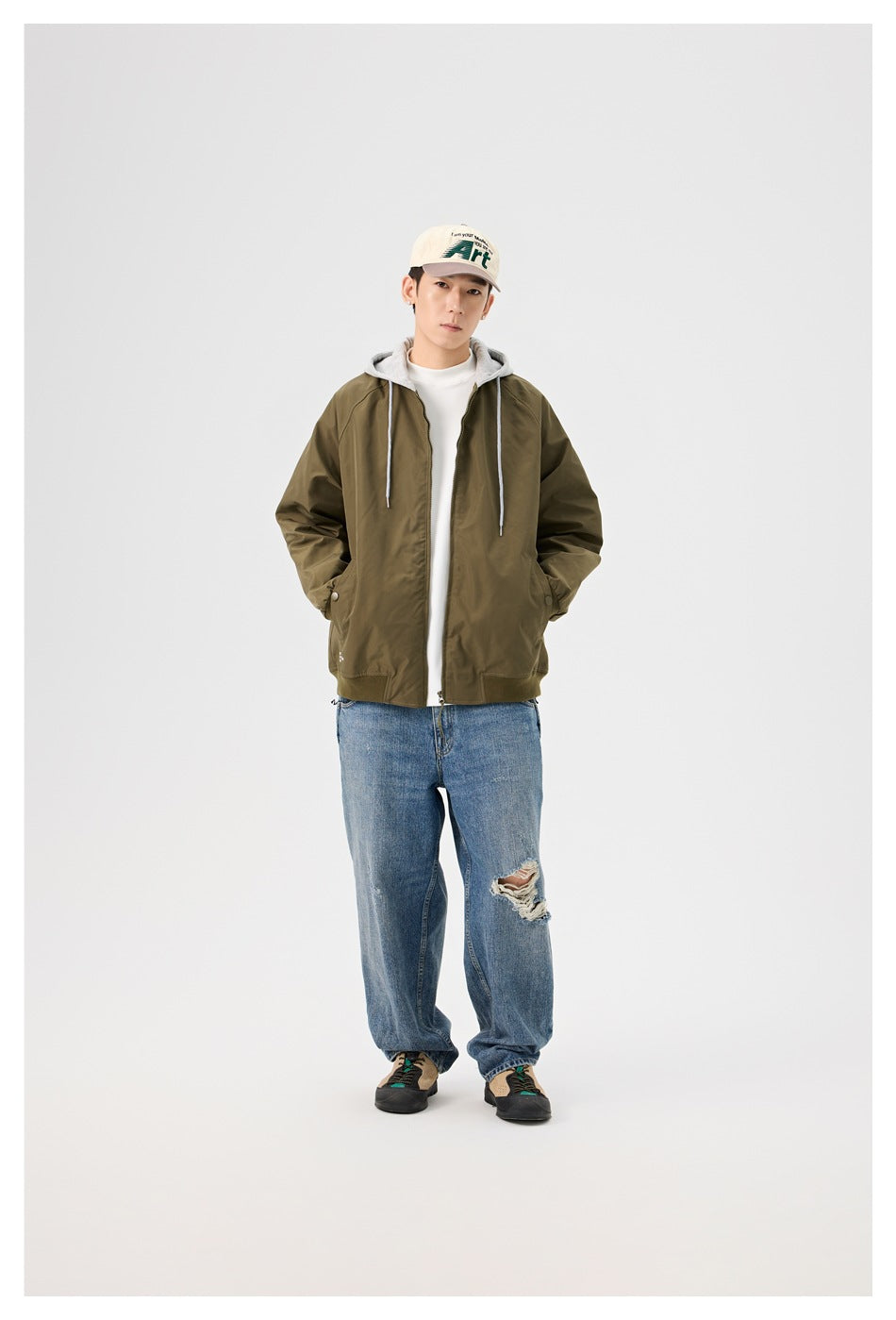 Model wearing the army green hooded zip jacket open over a white tee, with blue jeans, a cap and sneakers