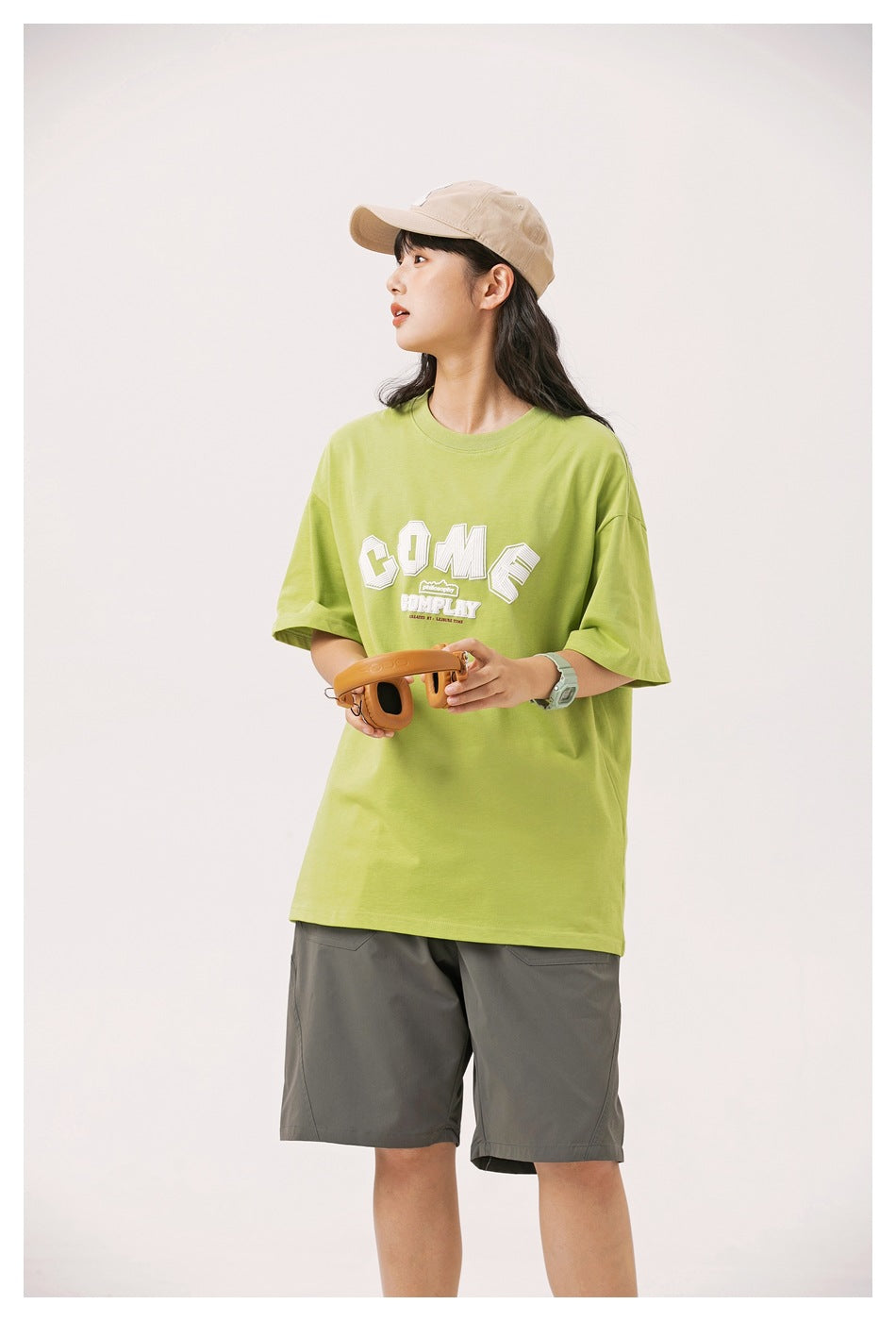 Model in a lime green graphic T-shirt and gray shorts, holding headphones in a three-quarter front view.