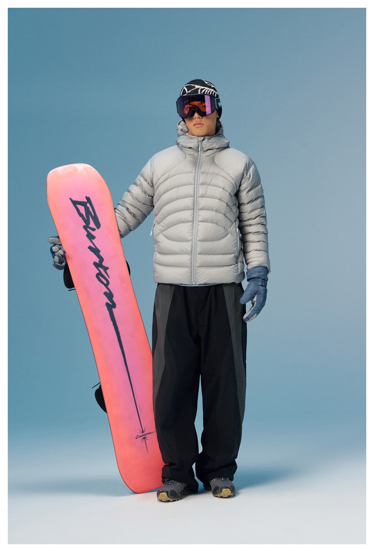 Man wearing a light gray quilted hooded puffer jacket and holding a pink snowboard.