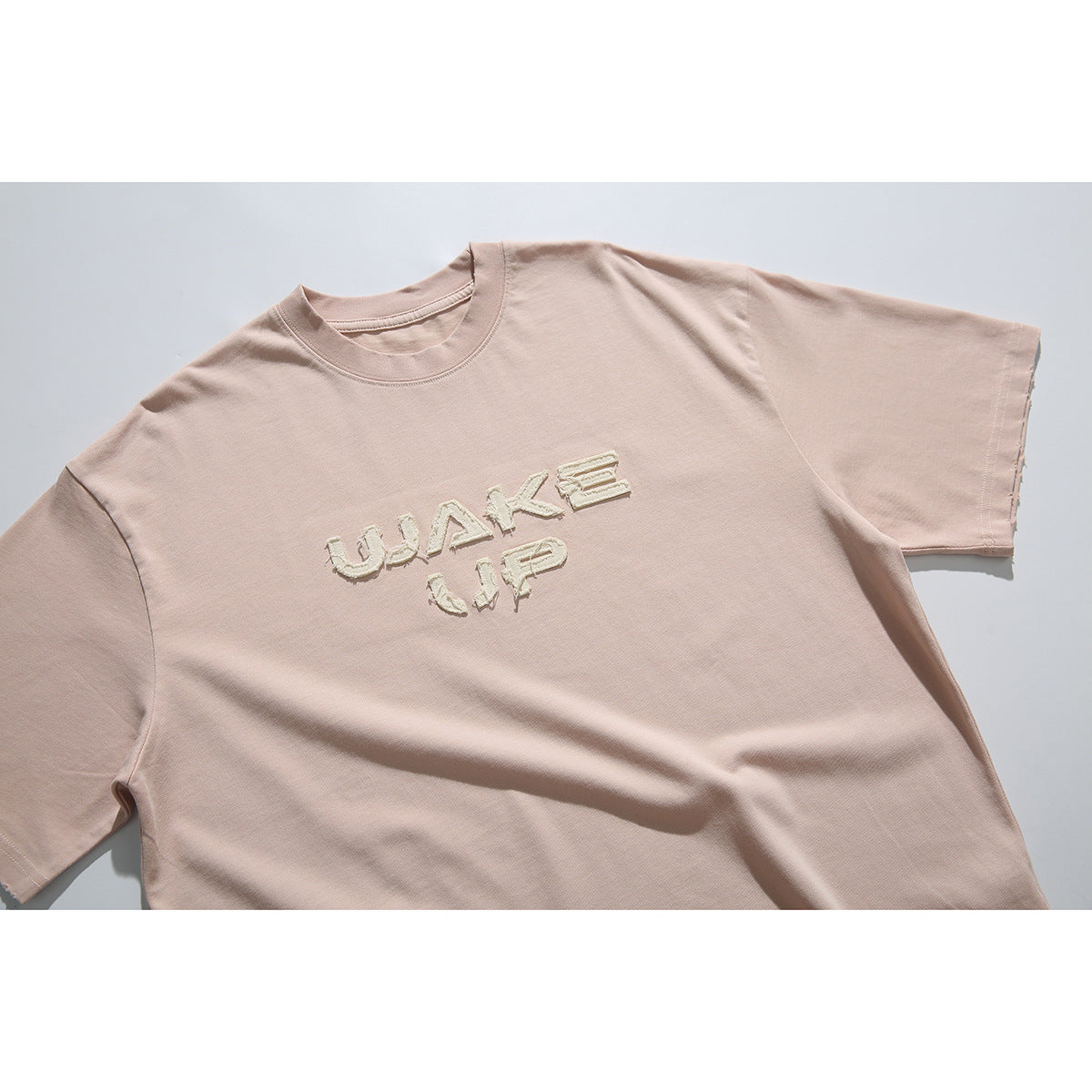 Close view of a pale blush-pink crew-neck T-shirt with cream textured “WAKE UP” lettering on the front.